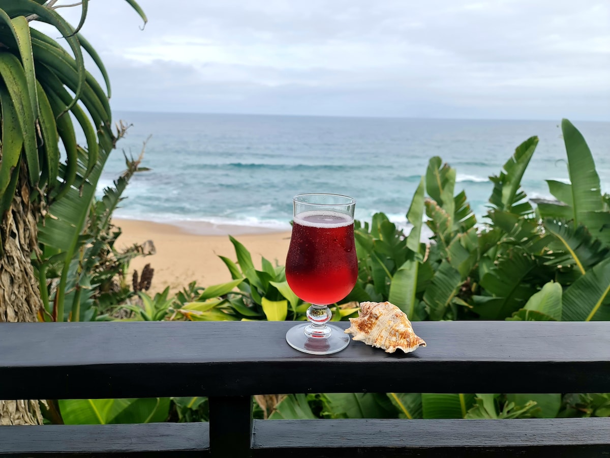 A refreshing drink is displayed on a wooden railing, accompanied by a seashell. Lush green foliage frames the scene, while the ocean waves gently lap the sandy shoreline in the background under a cloudy sky.