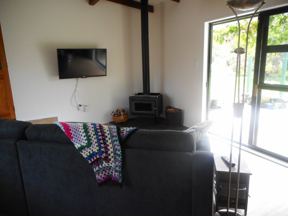 A cozy lounge area presents a dark gray sofa adorned with a colorful crocheted blanket. A modern wood stove and a wall-mounted TV are visible, with large glass doors allowing natural light to flow in from the outdoor space.