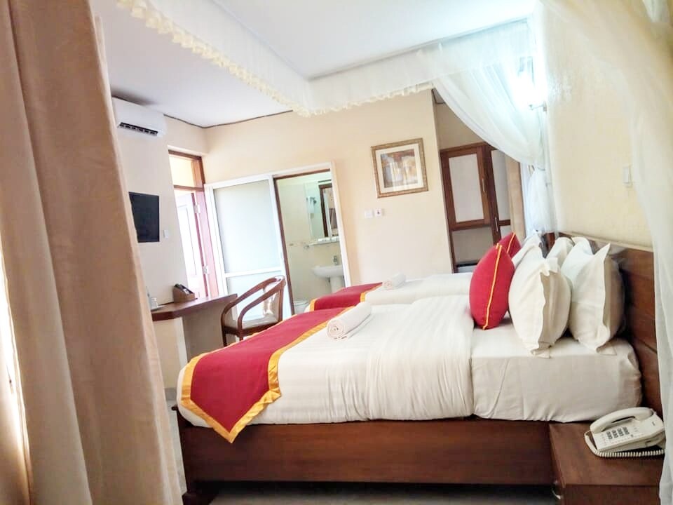 A room features two spacious beds adorned with white linens and decorative red accents. Soft lighting brightens the space, where a work desk and chair are positioned nearby. The room includes an attached bathroom, visible through an open doorway to the left.
