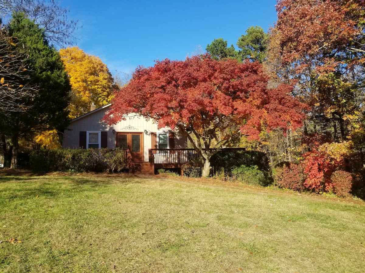A charming two-story home is framed by vibrant fall foliage, showcasing a deep red tree and lush greenery. The front porch invites guests with rustic wooden railings, while a spacious lawn provides plenty of room for outdoor activities.