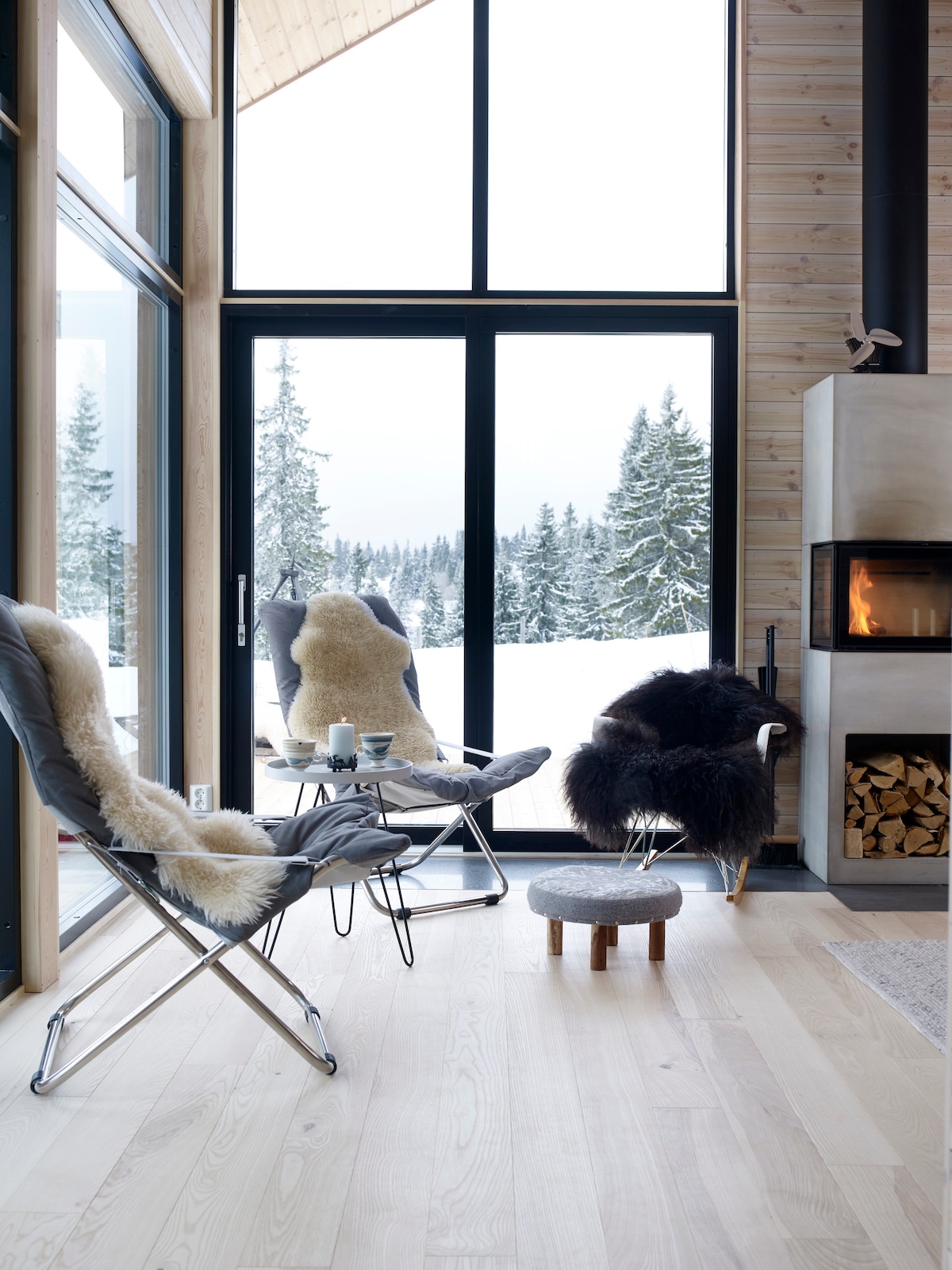 A bright and airy living space features two stylish chairs draped with faux fur throws. A cozy wood stove warms the room, while large windows display a snowy forest landscape outside. A small, round table sits nearby, enhancing the inviting atmosphere.