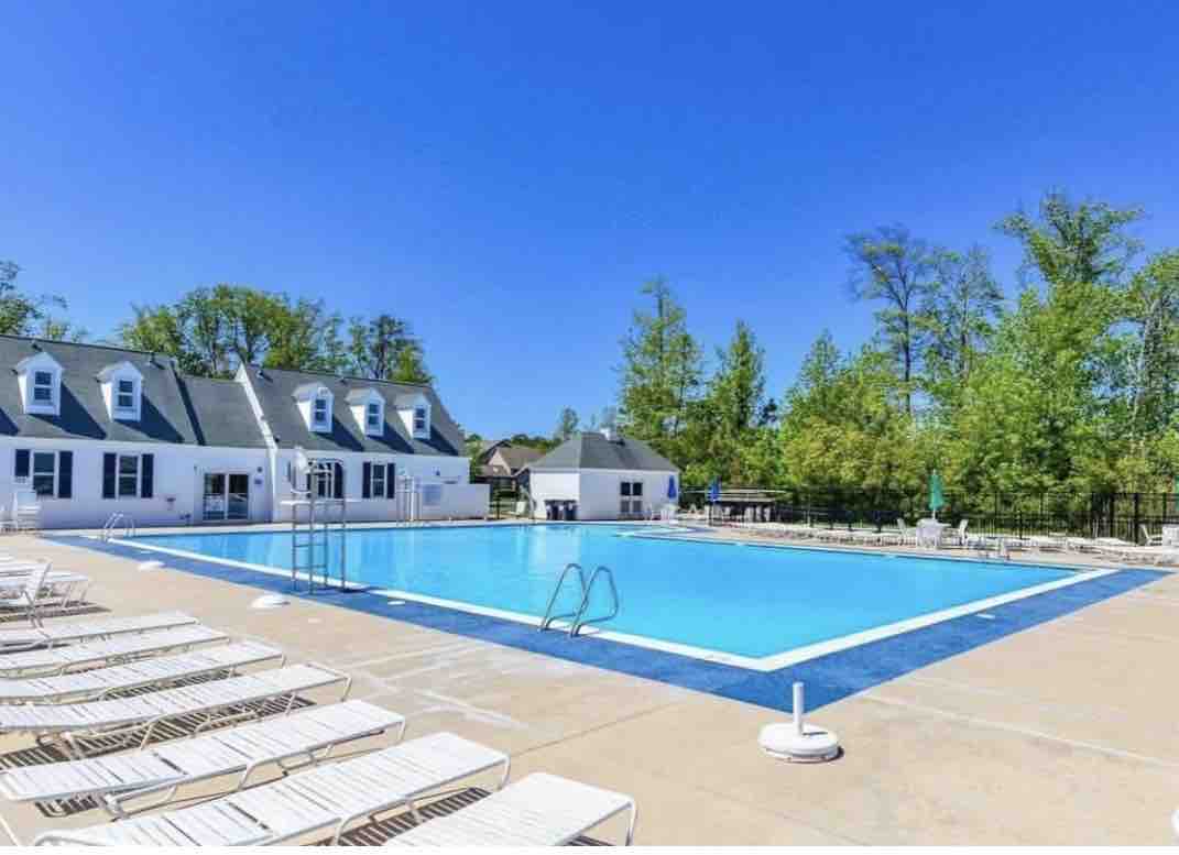 A large outdoor swimming pool is surrounded by a paved deck equipped with numerous lounge chairs for relaxation. Lush greenery frames the area, and a few trees provide a touch of shade. A white building is visible nearby, adding to the inviting atmosphere.