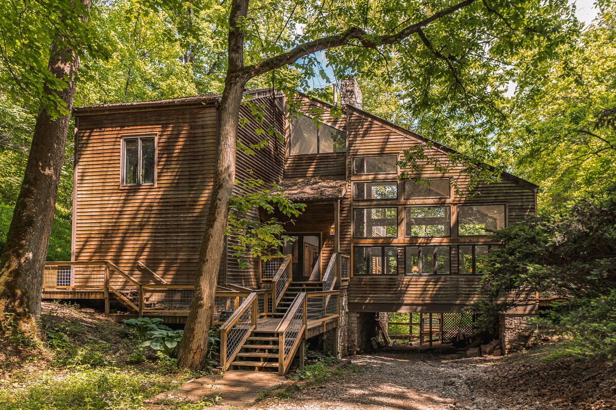 A large lodge nestled among trees features a wooden exterior with expansive windows that invite natural light. A sturdy staircase leads to a welcoming entryway, while the surrounding landscape includes a mix of vibrant green foliage, enhancing the connection with nature.