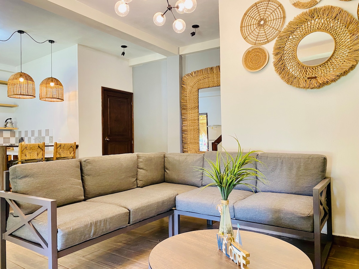 A spacious living area features a large sectional sofa in neutral tones, complemented by a round coffee table. Decorative woven mirrors adorn the walls, while pendant lights provide soft illumination. The open layout connects to the dining area and kitchen, enhancing the flow of the space.