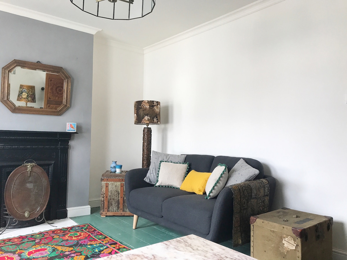 A cozy living area features a dark sofa adorned with a mix of decorative pillows. A rustic coffee table sits nearby, and an ornate lamp adds warmth. The wall displays a vintage mirror, while a colorful rug adds a soft touch to the floor.