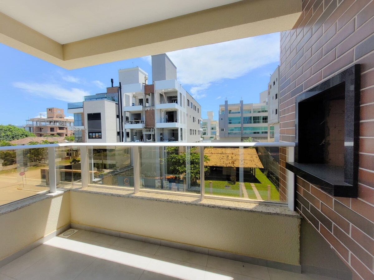 A spacious balcony features a built-in grill, surrounded by a glass railing. The view includes neighboring buildings and greenery below, with clear blue skies creating a bright atmosphere.
