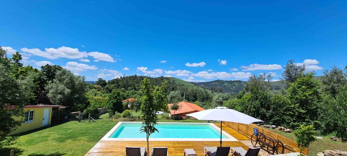 A serene outdoor area features a rectangular swimming pool surrounded by wooden decking. Sun loungers are positioned under a large umbrella, offering shade. Lush greenery and hills are visible in the background, adding to the peaceful setting. A few houses can be seen further in the distance.