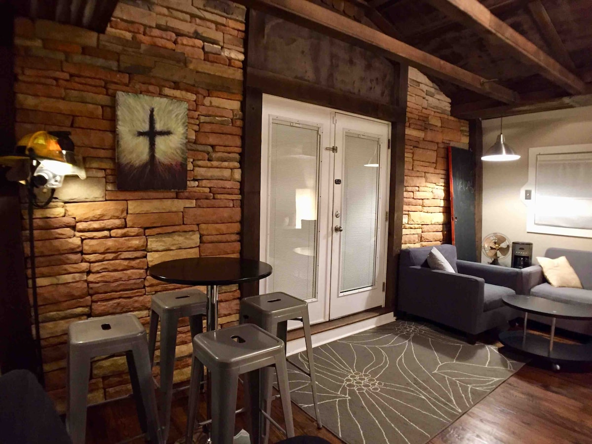 A cozy living area features textured stone walls and rustic wooden beams. A modern grey couch is positioned next to a circular coffee table, while metal stools offer additional seating. Large double doors provide natural light and access to the outside.