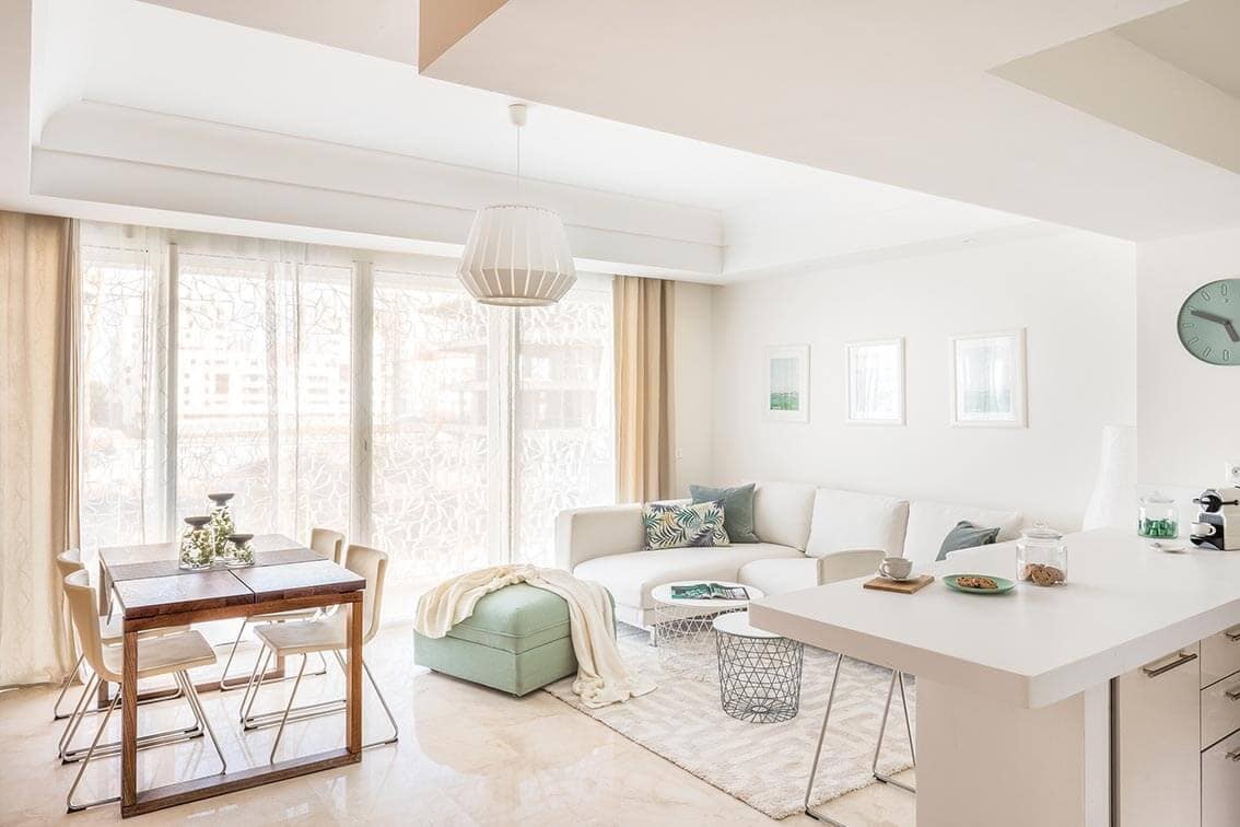 A bright living area features a comfortable sofa accompanied by decorative pillows, a light-colored rug, and a minimalist coffee table. A dining table with chairs sits to the side, while large windows allow natural light to fill the space. Simple art prints adorn the walls.