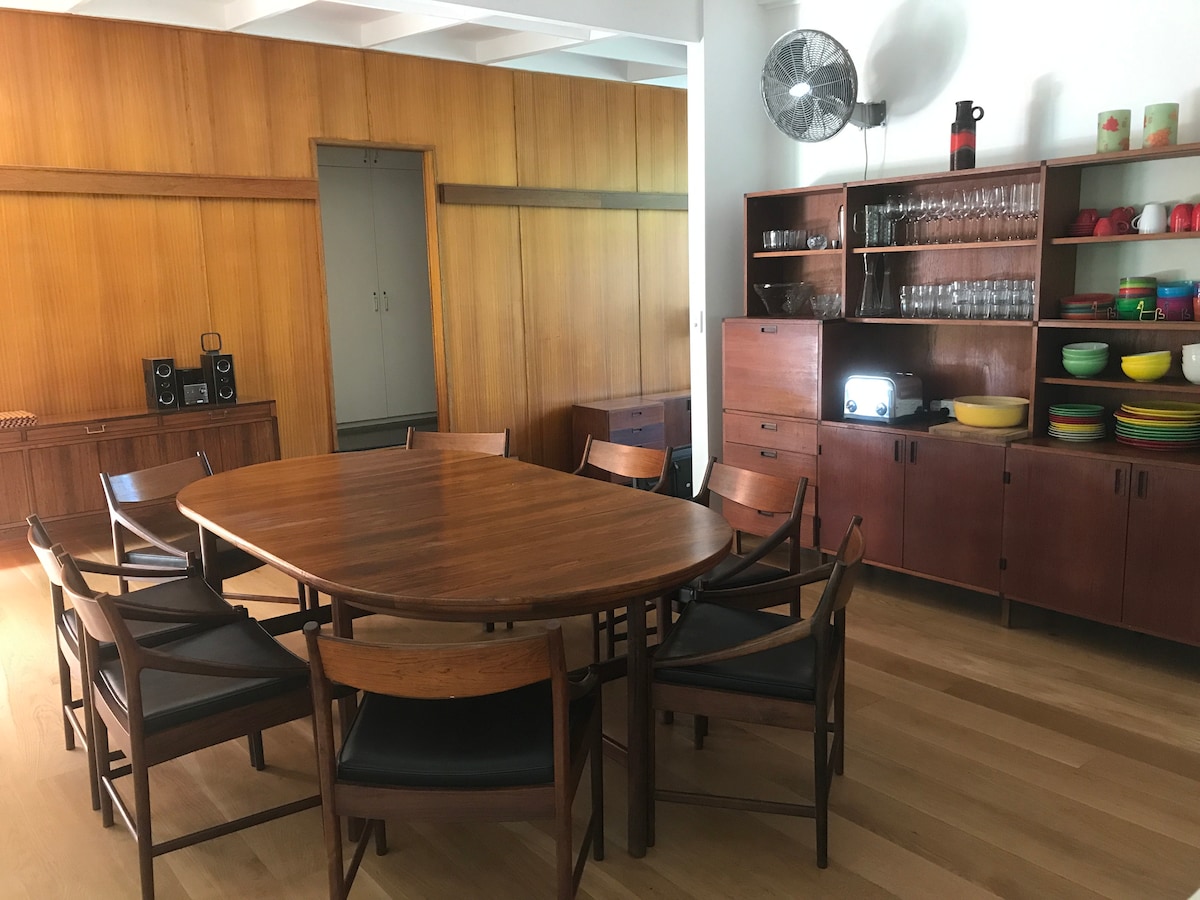 A mid-century dining area features a large round wooden table surrounded by eight wooden chairs. The walls are adorned with warm-toned wood paneling, complemented by a sideboard displaying colorful tableware and glassware. Natural light illuminates the space, contributing to a welcoming atmosphere.
