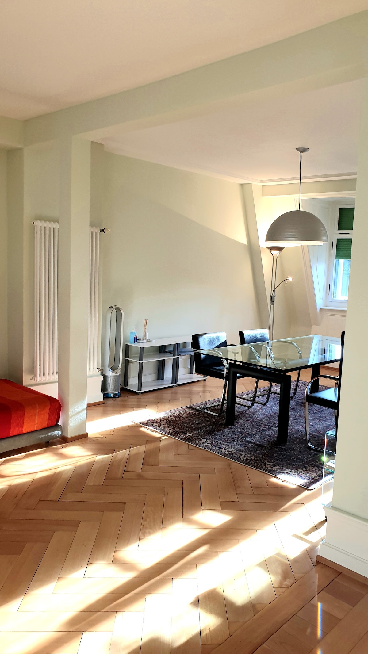 A light-filled dining area features a glass table surrounded by four black chairs. A sleek floor lamp stands beside a colorful couch, enhancing the modern design. Sunlight casts gentle patterns on the polished wooden floor, which exhibits a classic herringbone pattern.