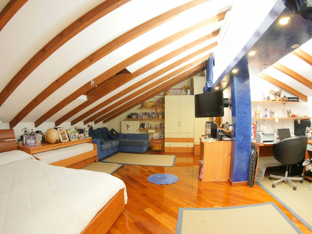 An attic room features a spacious layout with exposed wooden beams and a sloped ceiling. A large bed is positioned next to a cozy blue sofa. A computer desk is located in one corner, with shelves displaying books and decorative items.