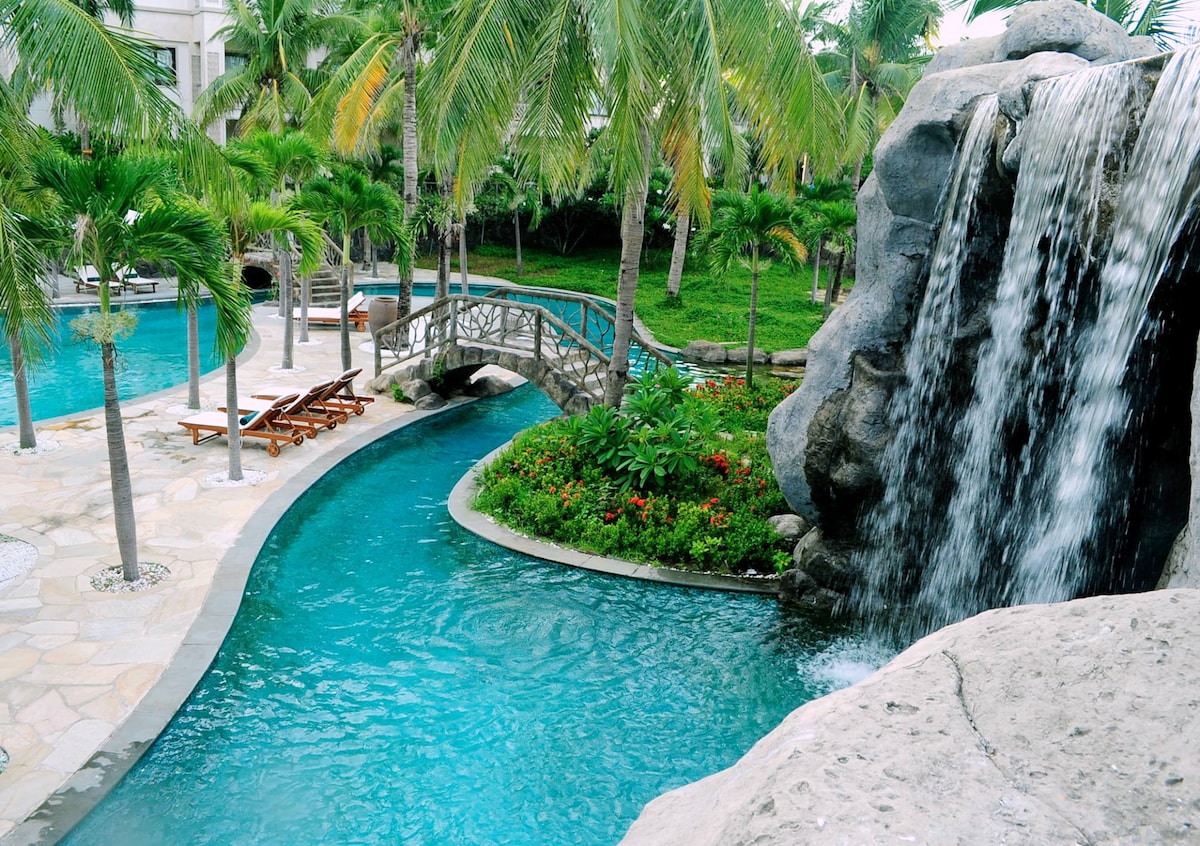 A serene pool area is depicted, featuring a waterfall cascading over a rocky formation into a clear blue pool. A stone bridge crosses the water, surrounded by lush tropical plants and palm trees, creating a refreshing and tranquil atmosphere.
