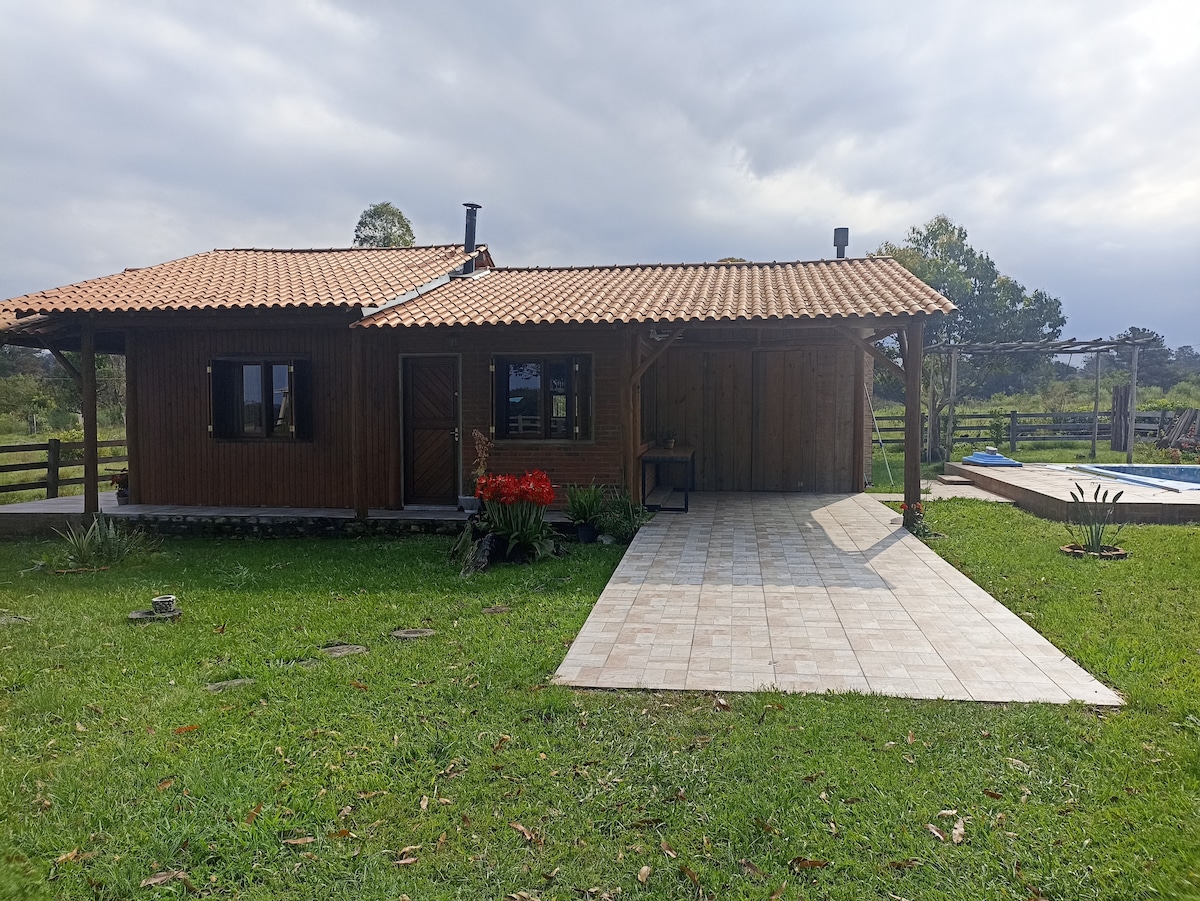 A charming wooden cottage features a tiled patio surrounded by lush green grass. The sloped roof is adorned with traditional tiles, and large windows allow natural light to fill the interior. A pool is visible in the background, framed by greenery.