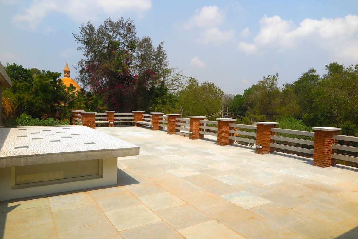 A spacious terrace is shown with a smooth stone floor and decorative brick walls. Surrounding trees and plants provide a natural setting. A unique structure with a conical roof is visible in the distance, complementing the serene outdoor space.