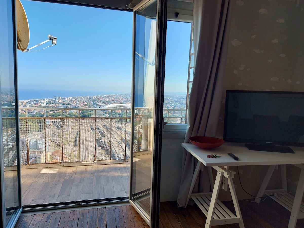 A spacious room features large glass doors leading to a balcony with panoramic views of the coastline and the city. A flat-screen television sits on a simple white table, while the wooden floor adds warmth to the modern decor.