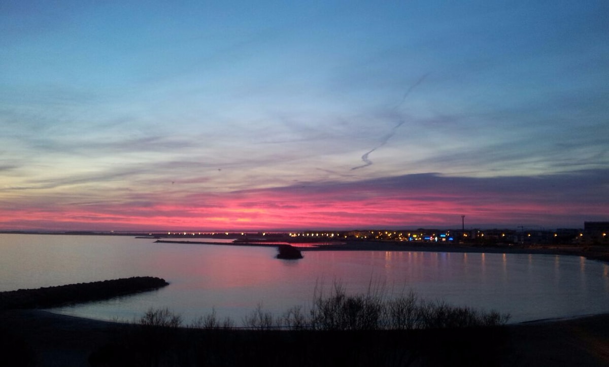 A serene view of a coastal landscape at sunset, featuring vibrant pink and purple hues in the sky. The water reflects these colors and hints of shoreline and lights from nearby buildings create a calm atmosphere along the beach.