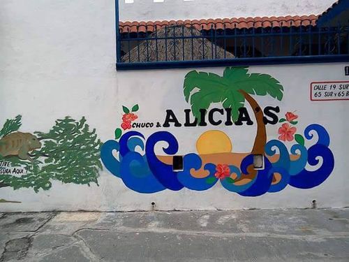 A vibrant mural features waves in blue hues, a sunset with an orange sun, and palm trees. The name 'Chuco & Alicia's' is prominently displayed, surrounded by tropical flowers. To the left, a green bush with a playful iguana is depicted.