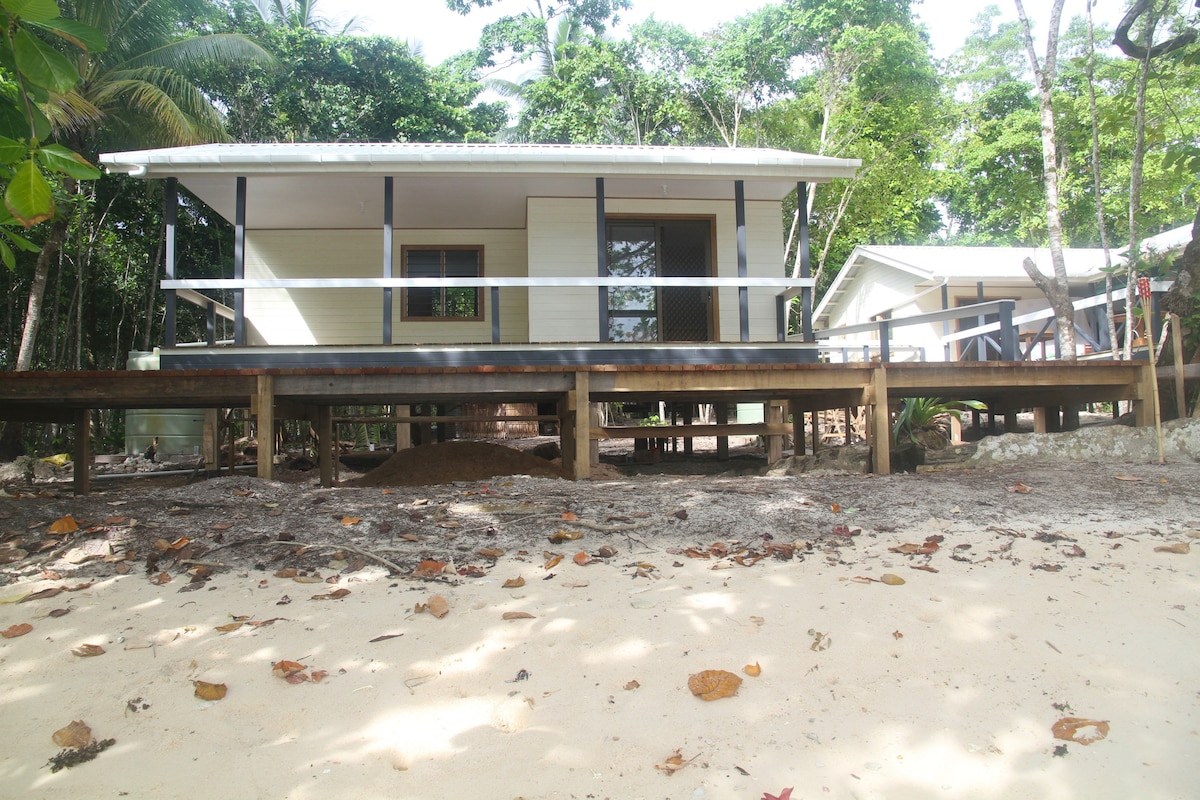 A bungalow with a white facade is set against lush greenery, showcasing a large deck with wooden railing. The sandy area in front of the bungalow features scattered leaves, and a gentle slope leads to the surrounding nature.