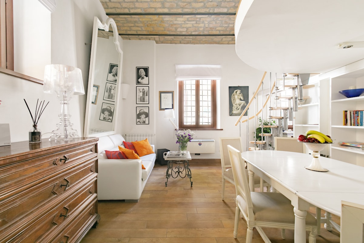 The living area features a light wood floor accented by a white sofa adorned with colorful cushions. A wooden dresser adds warmth, while framed photos and artwork enrich the walls. A dining table with white chairs is set near a staircase leading to the mezzanine bedroom.