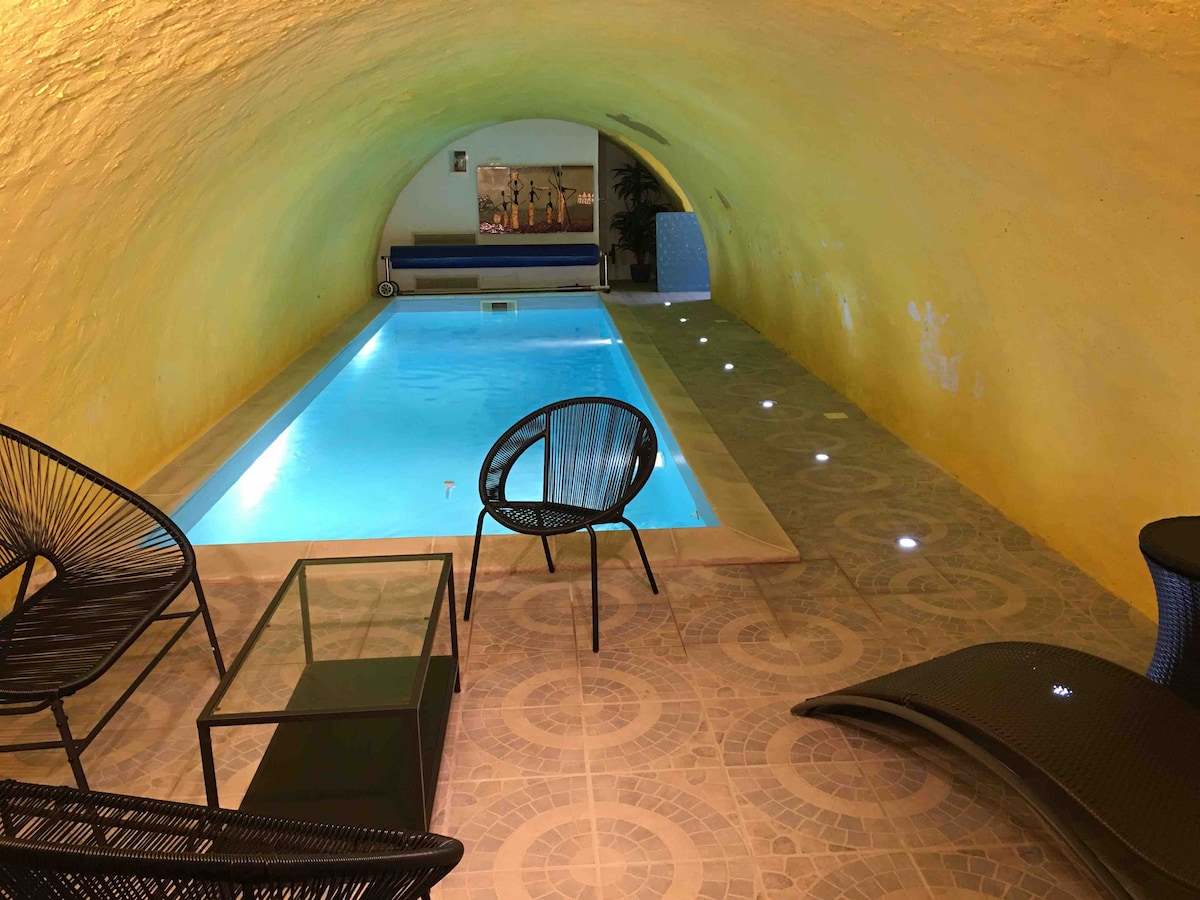 An indoor swimming pool is highlighted in a vaulted, yellow-hued space. Two black chairs and a glass table accompany the pool area, surrounded by warm lighting that enhances the serene environment. A blue cushioned bench is positioned at one end, offering additional seating.
