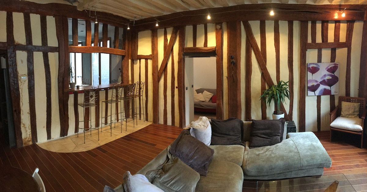The image depicts a living area featuring exposed wooden beams and a blend of modern and rustic decor. A spacious sectional sofa is arranged to invite relaxation, complemented by a small armchair. A bar area with high stools sits alongside the back wall, enhancing the open layout.