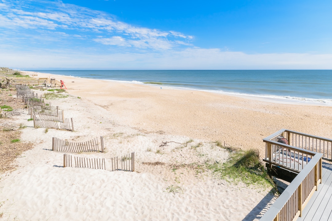An expansive view of a sandy beach stretches towards the horizon, where the ocean meets the sky. Gentle waves roll onto the shore, and sparse wooden fencing complements the natural landscape. A clear blue sky with scattered clouds creates a serene atmosphere.