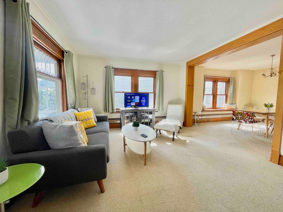 A bright living area is showcased, featuring a comfortable gray sofa accented with decorative pillows. A television is centered on a media console. Sunlight streams through large windows, illuminating the space. A dining area with a light-colored table and chairs is visible in the background.