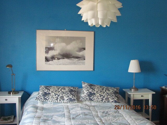 A comfortable sleeping area features a spacious bed with patterned pillows and a decorative quilt. Blue walls provide a vibrant backdrop, complemented by framed artwork depicting ocean waves. Accent lighting is provided by a modern lamp on one nightstand and a stylish pendant light overhead.