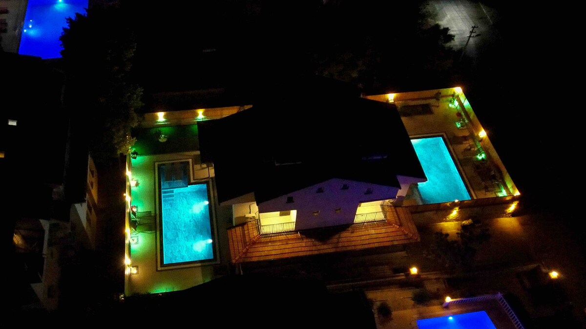 An aerial view captures a two-story villa surrounded by illuminated pool areas. The cooling turquoise water contrasts with the soft lighting around the outdoor spaces. Pathways are lined with lights, enhancing the welcoming ambiance of the evening setting.