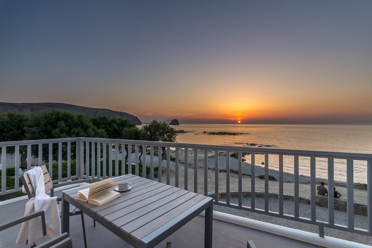 A balcony with wooden furnishings overlooks a serene coastal scene at sunset. The horizon is painted in warm hues as the sun descends, reflecting on the calm waters. The landscape includes rocky shorelines and lush greenery in the background.