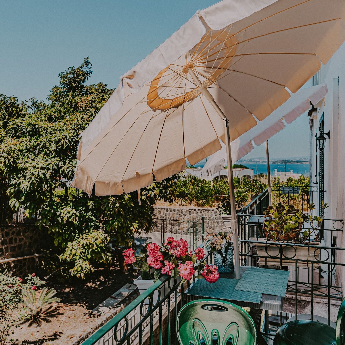 An outdoor area features a green table surrounded by chairs, shaded by a large umbrella. Potted pink flowers adorn the space, complemented by lush greenery in the background. A view of the sea can be glimpsed beyond the garden.