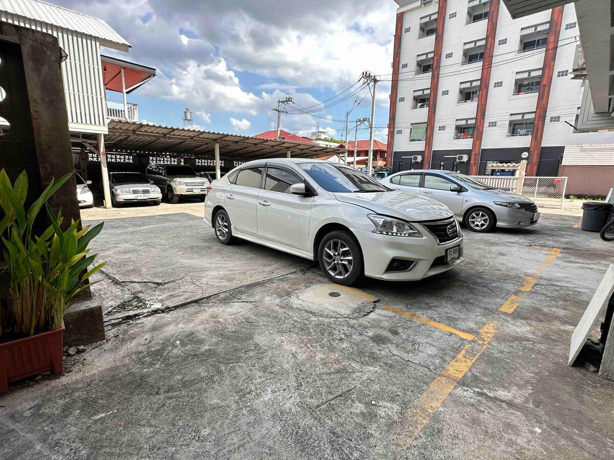 The image shows an outdoor parking area with several parked vehicles. A white car occupies a marked space, while another gray car is visible in the background. Surrounding greenery is present near the edge of the parking lot, providing a natural touch to the scene.