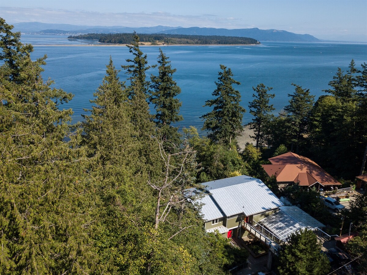 Aerial view showcasing the Salish House nestled among tall trees, with panoramic vistas of Hale Passage and the surrounding coastline. The tranquil blue water reflects the sky, while neighboring properties are visible in the green landscape.
