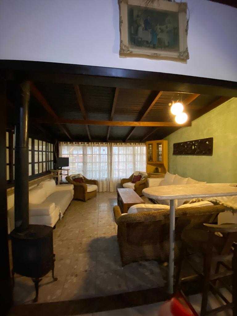 A cozy living area is showcased with a combination of white and natural-fiber seating. Warm lighting illuminates the space, enhancing the wooden ceiling details. Large windows are dressed with sheer curtains, allowing soft light to filter in, creating a welcoming environment.