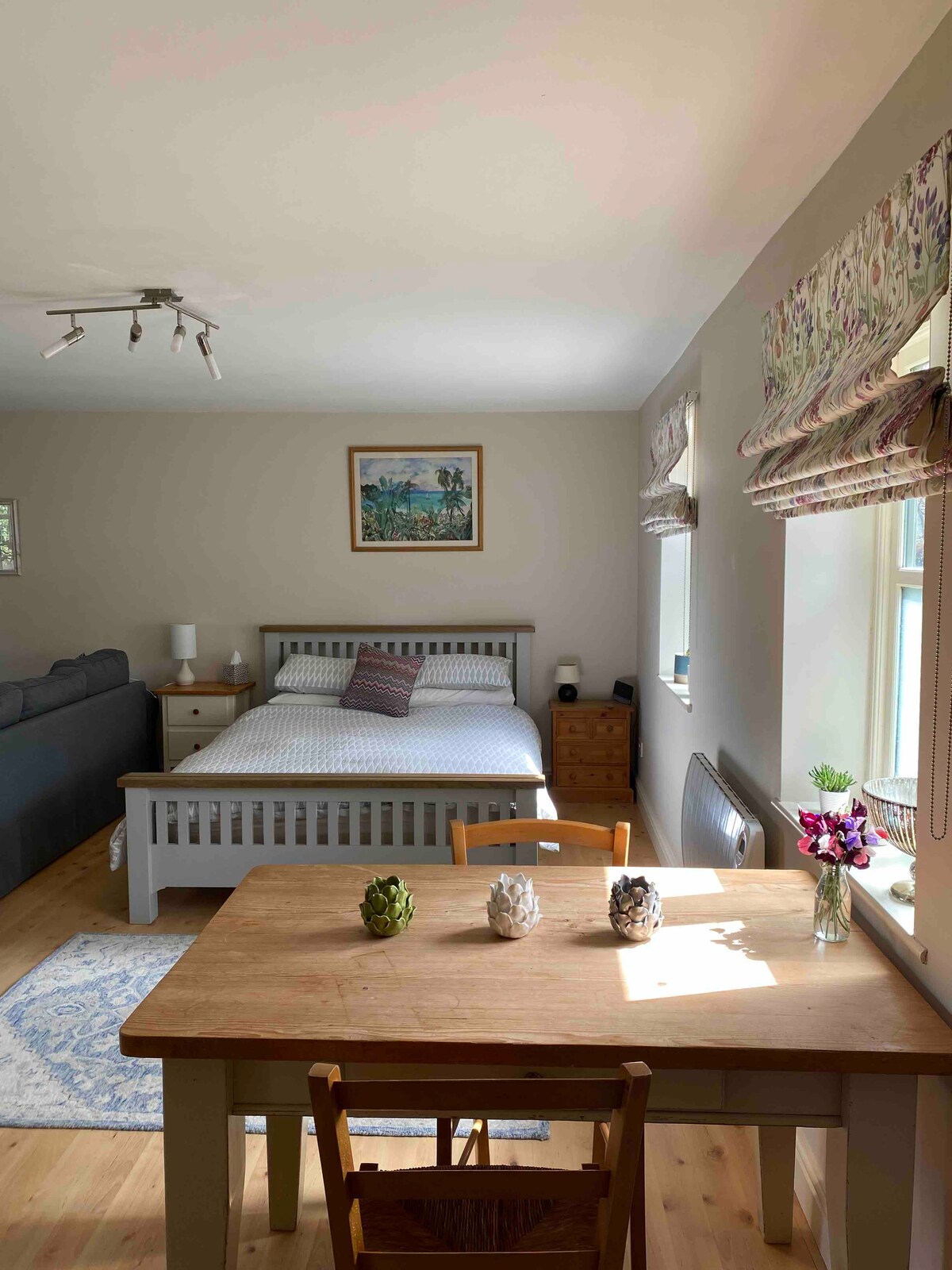A well-furnished studio features a spacious bed with a neutral-tone headboard, accompanied by bedside tables. A dining area with a wooden table and chairs is in view, while a sofa is positioned in the background. Natural light fills the room through two windows adorned with patterned shades.