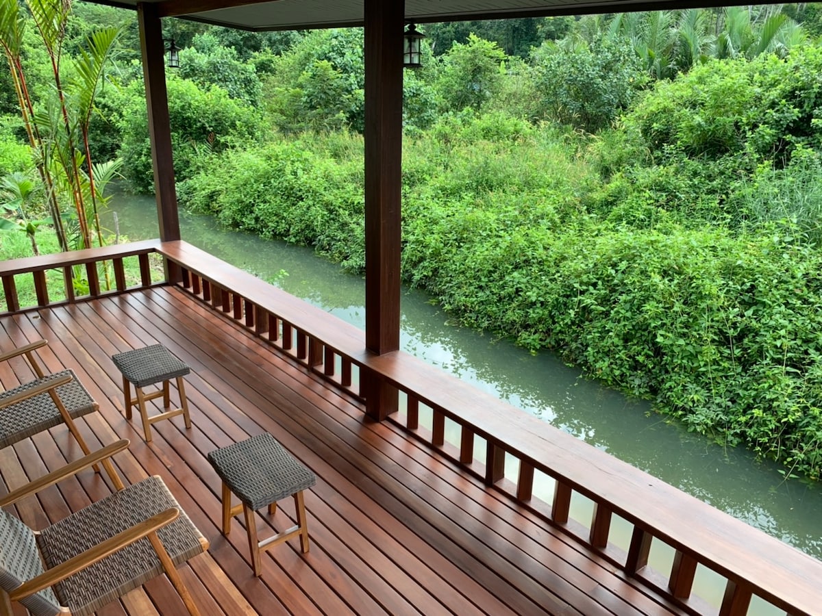 A spacious balcony is featured, overlooking a tranquil canal bordered by lush greenery. Four wooden chairs are positioned for comfort, while the smooth wooden deck complements the natural surroundings.