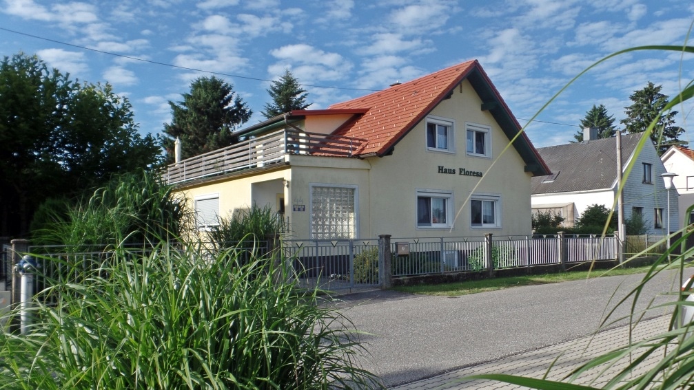 A two-story yellow house with a red roof is framed by lush greenery. The structure features a balcony on the upper level and multiple windows, while a fence surrounds the property. The clear blue sky and scattered clouds provide a calm backdrop.