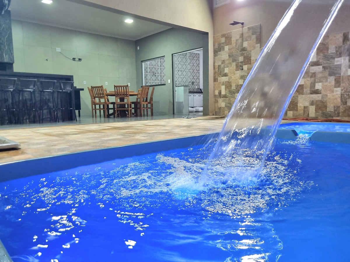 A heated pool features a waterfall, creating gentle ripples in the vibrant blue water. In the background, a dining area with a wooden table and chairs is visible, surrounded by a well-lit indoor space accented by natural stone tiles.