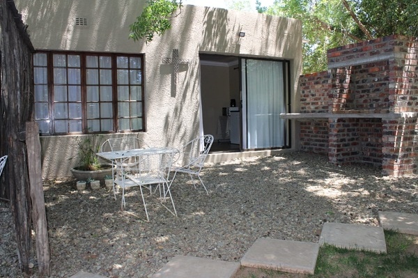 A gravel patio area is shown, featuring a small round table with four white chairs. A private braai area made of brick is positioned nearby, with a large opening leading into a kitchenette visible through glass sliding doors. Natural light filters through the trees.