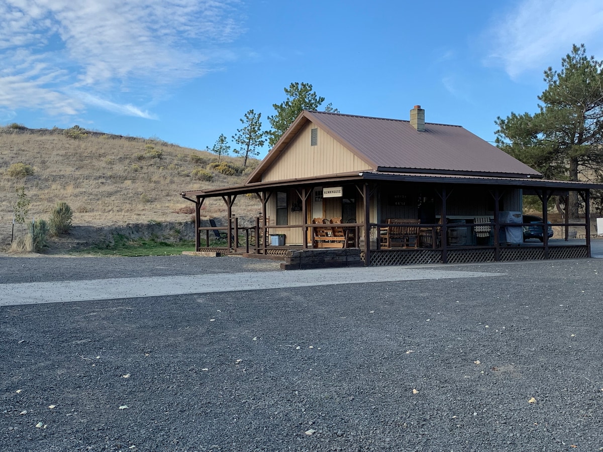 A spacious bunkhouse features a covered porch and rustic wooden furnishings, nestled against sloping hills. The gravel drive leads to the entrance, framed by natural vegetation and clear blue skies.