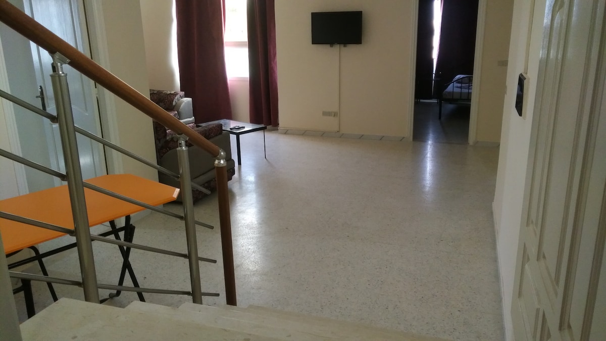 A spacious entryway featuring a decorative staircase with metal railing. A small orange table is positioned near a comfortable seating area with a patterned armchair. In the background, a television is mounted on the wall, and an additional room is visible, adding depth to the space.