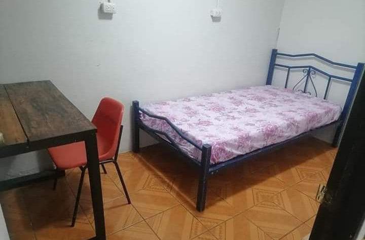 A simple room features a single bed with floral bedding and a wooden table complemented by a red chair. The floor is tiled, and the walls are painted white, creating a clean and functional space.