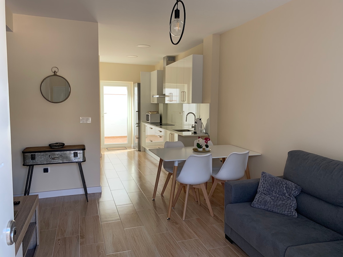 A modern kitchen and dining area are visible, featuring light wood cabinetry and a white table with four chairs. A gray sofa sits to one side, while a round mirror is mounted on the wall. The space is filled with natural light through an open doorway.