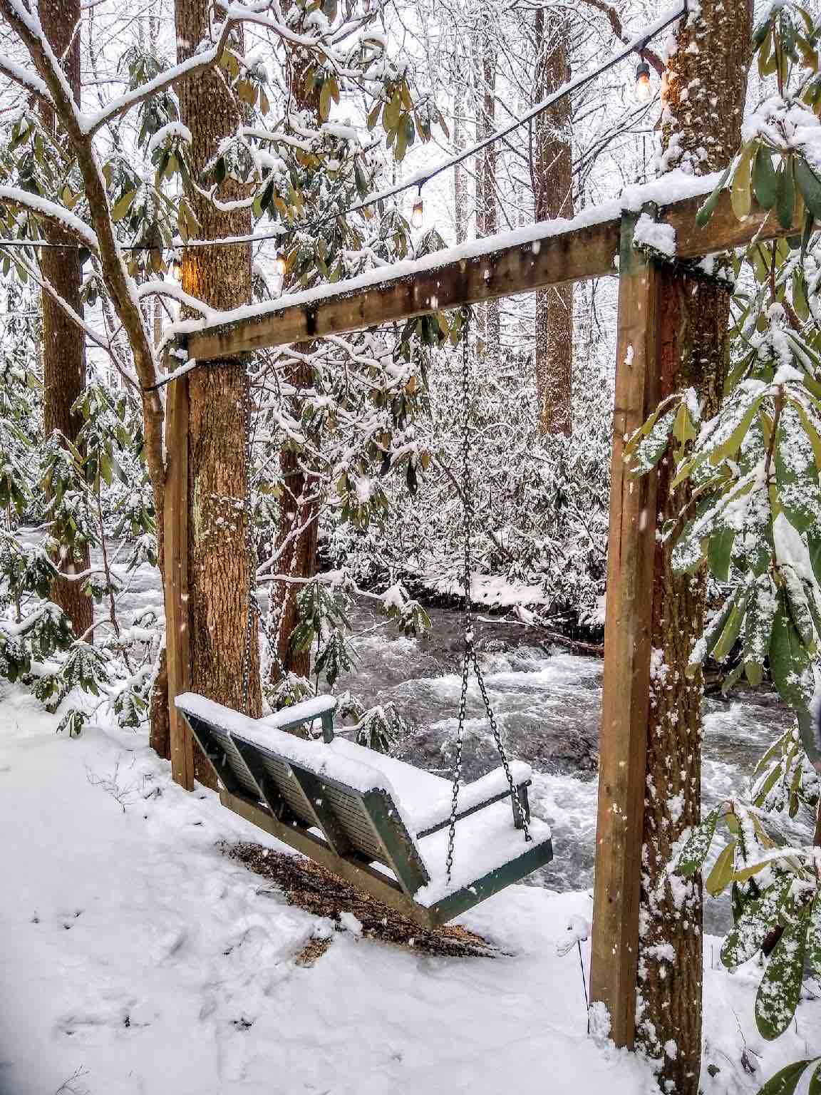 A wooden swing hangs from a sturdy frame, surrounded by snowy landscapes and evergreen trees. The creek flows gently nearby, its water partially visible through the snow-covered branches, creating a serene winter scene.