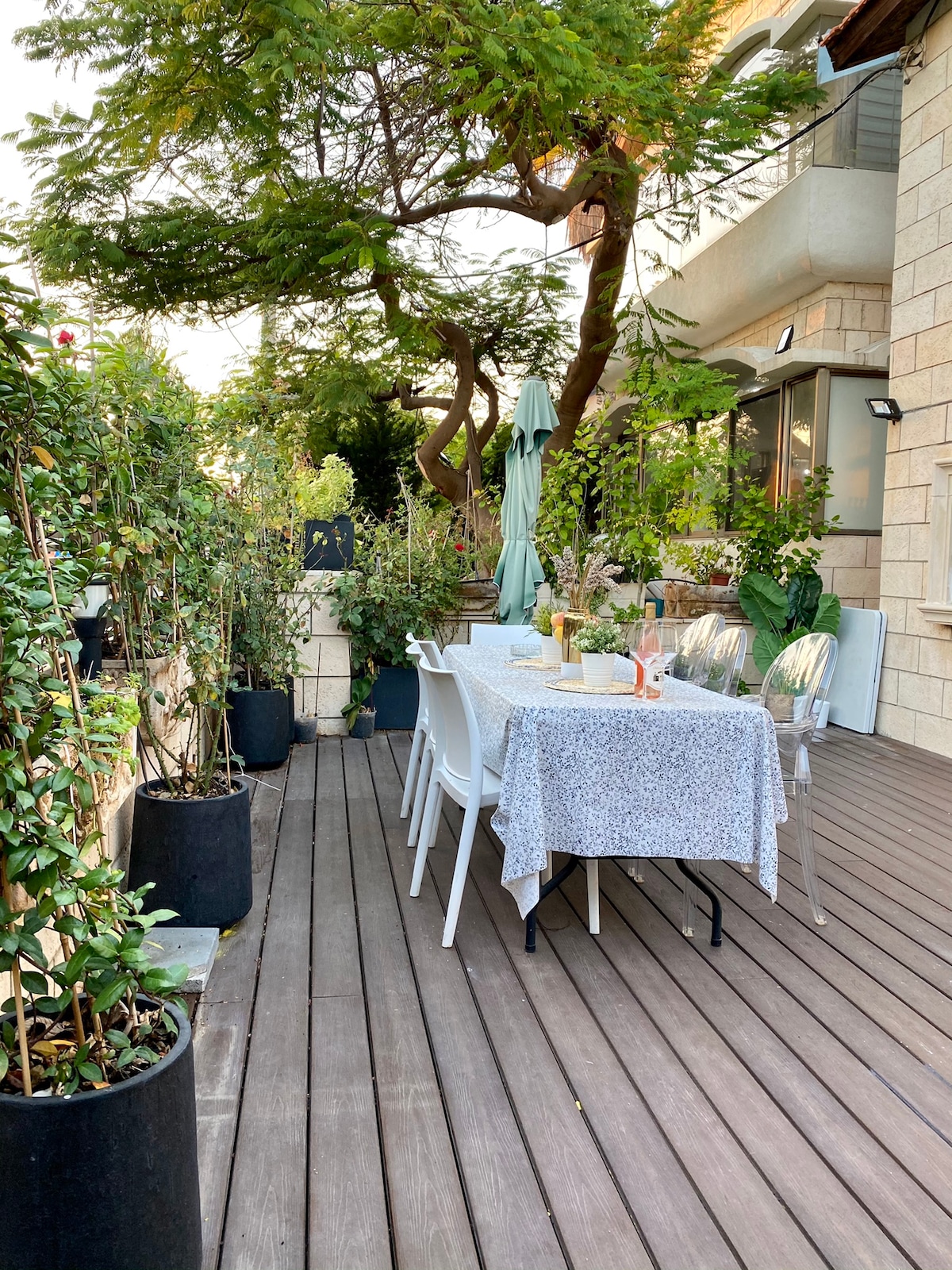An outdoor dining area is set with a table covered in a light tablecloth, surrounded by chairs. Green plants and trees create a lush backdrop, enhancing the sense of privacy. Sunlight filters through the leaves, casting soft shadows on the wooden deck.