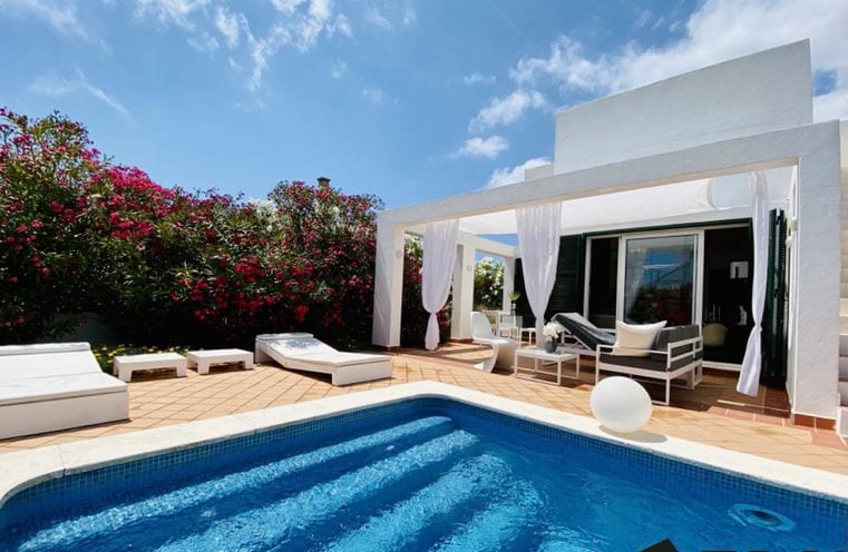 An outdoor area surrounds a lovely pool, with lounge chairs positioned nearby. Bright flowers enhance the vibrant environment. A shaded terrace is visible, offering seating for relaxation and enjoyment. The clear sky reflects on the water, completing the inviting space.