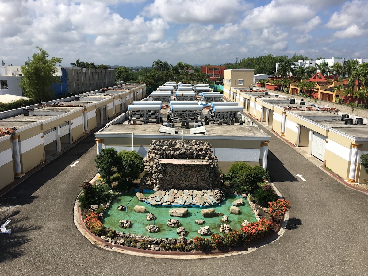 An aerial view reveals a well-maintained property layout featuring multiple buildings arranged around a central landscaped area. A water feature comprised of rocks and plants is visible, with colorful flora lining the pathways and enhancing the serene atmosphere.