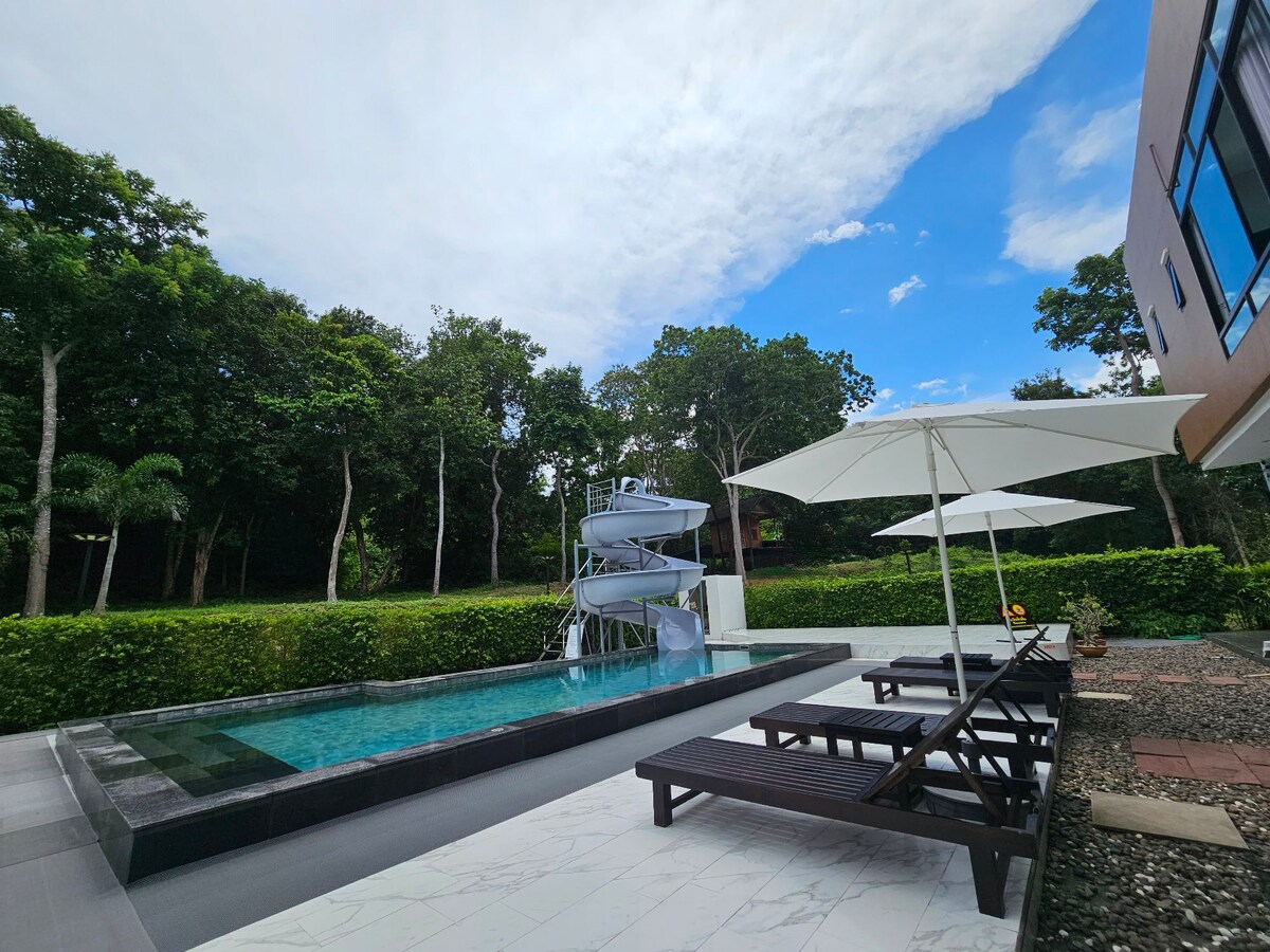 An outdoor area features a sparkling pool accompanied by a water slide, surrounded by lush greenery. Comfortable lounge chairs are positioned near the pool, with large umbrellas providing shade. The blue sky complements the tranquil setting, enhancing the inviting space.