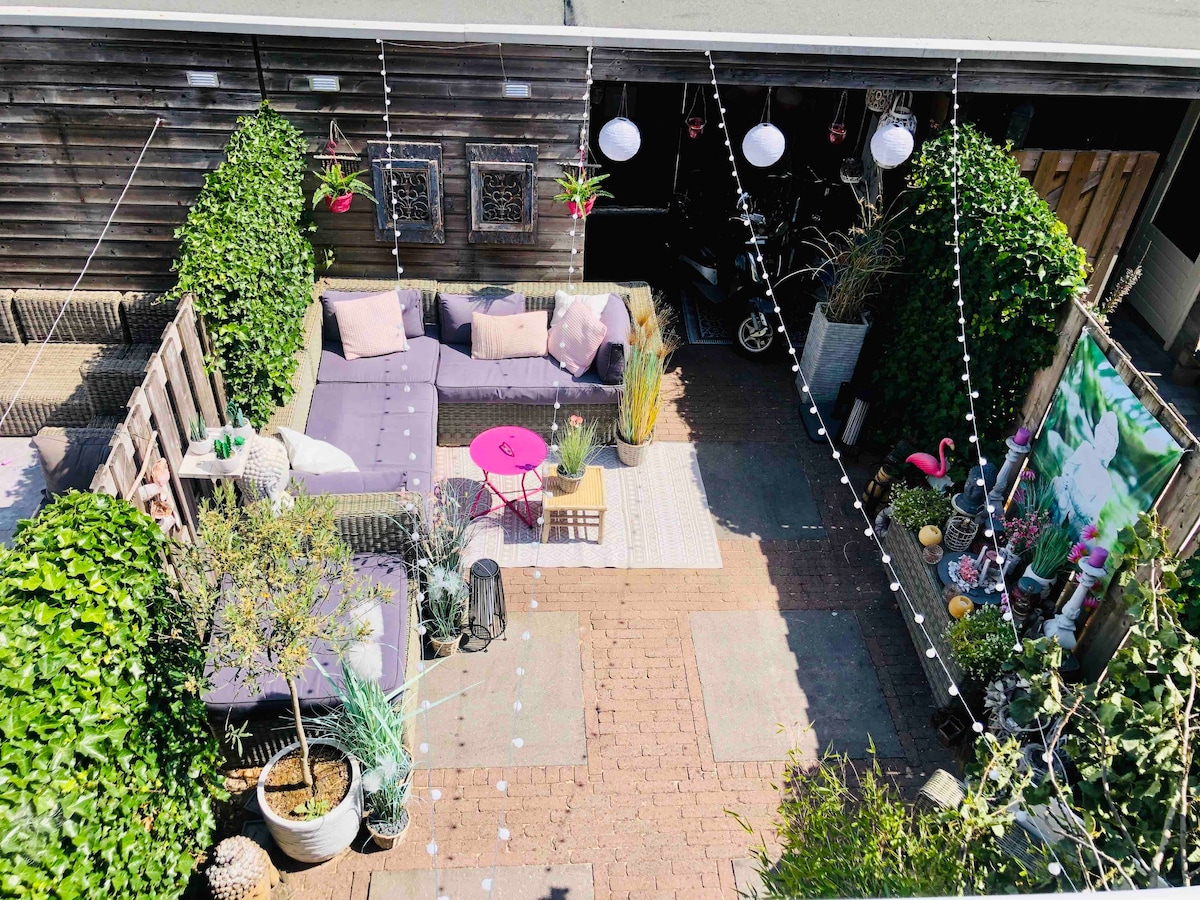 An outdoor area is highlighted with a large purple sectional sofa surrounded by lush greenery and potted plants. A small pink table adds a pop of color, while string lights and lanterns provide soft illumination. Decorative elements include a wall-mounted artwork and various garden ornaments.